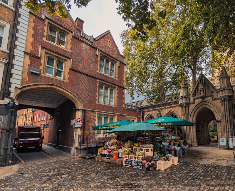 A vibrant flower stall located on a cobblestone street in front of historic red-brick buildings with arched entrances and medieval architectural details, including decorative stonework and pointed arc