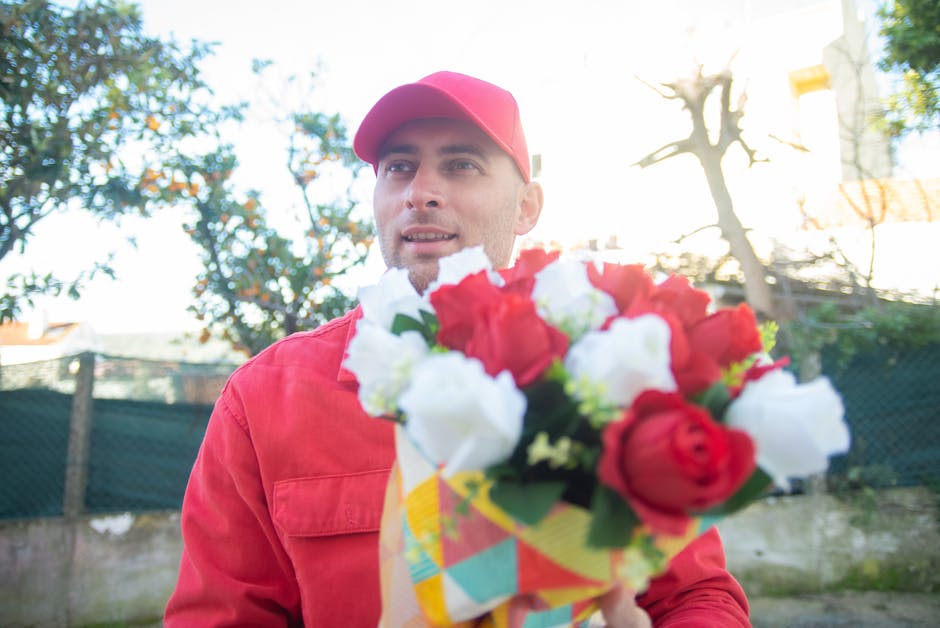 A cheerful delivery person dressed in a red uniform and matching cap is holding a vibrant bouquet of fresh roses with red, white, and pink petals, wrapped in colorful geometric-patterned paper. The bo