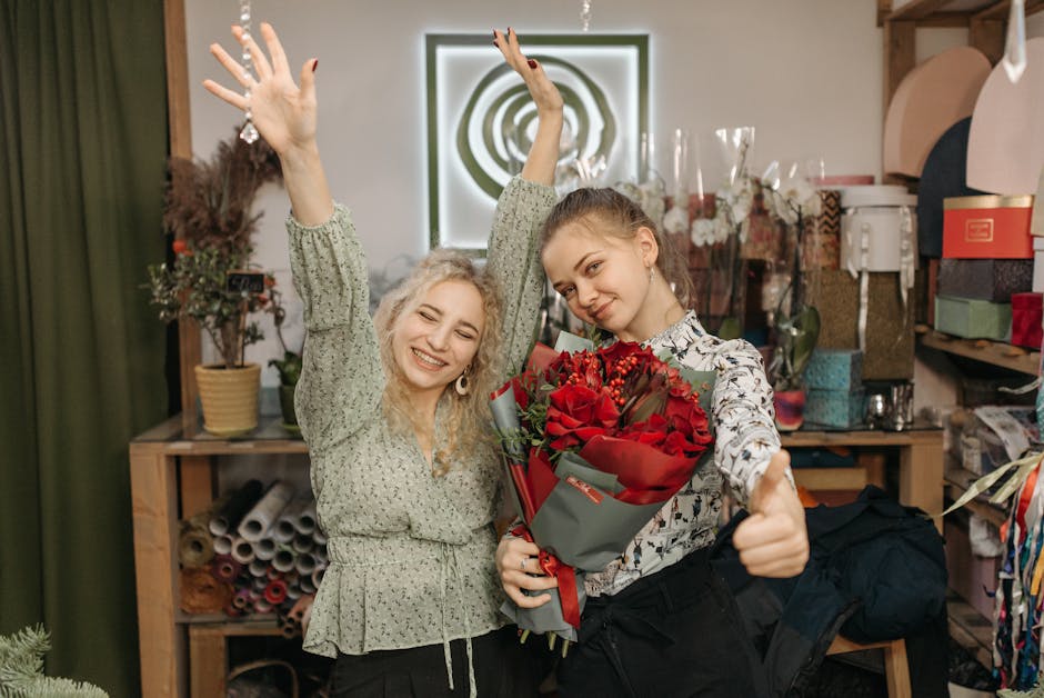 Two women are inside a florist shop, with one holding a vibrant bouquet of red roses wrapped in gray and red paper, while the other is smiling broadly with her arms raised in celebration. The arrangem