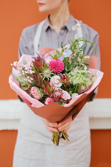 A person dressed in a light grey shirt and white apron, partially visible from the shoulders down, holding a large, vibrant bouquet of fresh flowers. The bouquet includes pink and white blooms such as