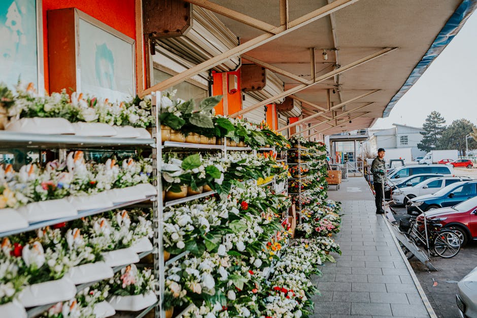 A colorful outdoor flower market set up along Harrow Road near Harrow Town Centre, featuring multiple shelves filled with fresh floral arrangements and bouquets. The flowers include various types such