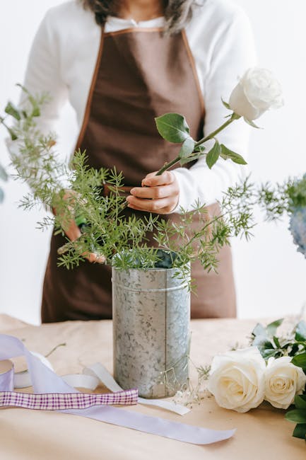 A florist wearing a brown apron over a white shirt arranging a floral bouquet with a single large white rose and green foliage, including delicate fern-like leaves, in a tall, rustic metal vase. The t