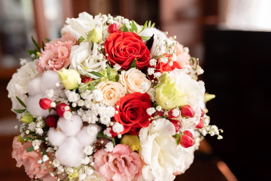 A bridal bouquet containing a mix of fresh flowers including vibrant red roses, soft pink roses, white lisianthus, and delicate baby's breath, arranged in a rounded, lush style. The bouquet is wrapped