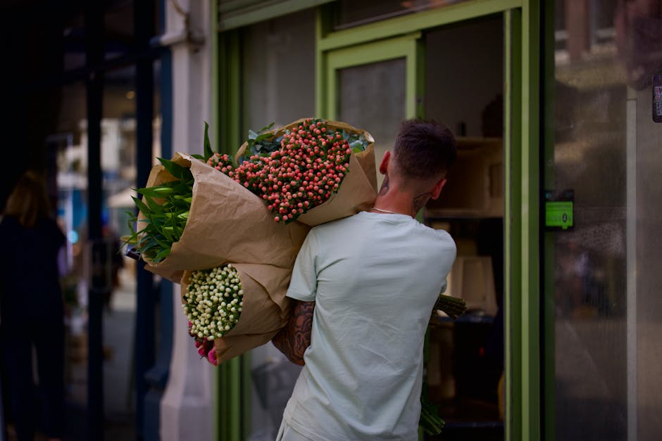 A young man with short dark hair, wearing a light-colored t-shirt, is holding a large bouquet of fresh flowers wrapped in brown paper outside a flower shop with bright green-framed windows. The bouque