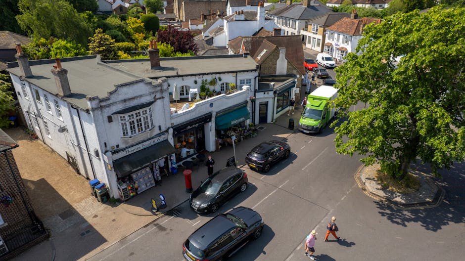 An aerial view of a small, white, two-story building with large windows and a green awning, situated on a street in Wealdstone HA3. The building houses a flower shop, as indicated by floral displays o